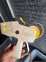 Close-up photo of handheld label gun with orange label roll inserted, showing condition and key features.