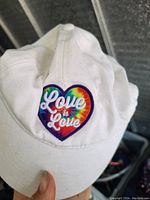 Photo shows front of white baseball cap with colorful heart patch and embroidered text 'Love is Love'. The cap is lightly used and held by a hand for scale.