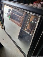 Large rectangular mirror with detailed carving on wooden frame, resting against wall in room scattered with other furniture