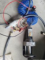 Photo of a black cylindrical pump with attached plumbing fittings, red and black wires, and a blue coiled hose with a valve knob, placed on a concrete floor.