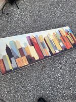 Framed rectangular painting showing an abstract city skyline with various colored buildings under natural light outdoors on gravel surface.