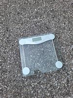 Digital glass bathroom scale placed on asphalt surface, visible rectangular platform and digital screen in white housing