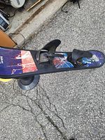 Photo showing the top side of a snowboard with attached bindings. The snowboard top has a multi-colored graphic design in red, blue, and purple. The bindings are black and secured on the board.