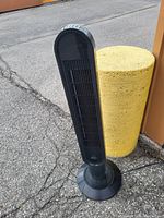 Black stand up tower fan outside near yellow bollard.