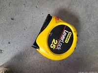 Yellow Stanley tape measure with Level Lock 25 ft label on black circular background on front of the case lying on a grey concrete surface.