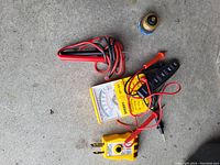 Five electrical related items placed on a concrete surface including an analog multimeter, outlet tester, wire stripper, test probes, and WD-40 spray can.