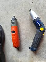 Photo shows two handheld cordless power tools on a concrete surface. The left tool is an orange Black & Decker 2.4V screwdriver. The right tool is a larger blue and gray tool with a yellow trigger, brand unknown.