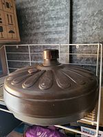 Photo displaying a large round, dark metal umbrella stand with embossed decorative pattern and central knob on top.