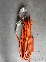 Photo of a handheld portable work light with a metal protective cage around the bulb, attached to a metal handle and a long orange power cord coiled up beside it.