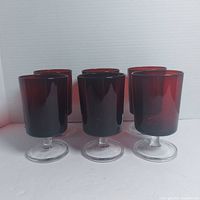 Six deep red glass goblets with clear stems and bases arranged in a group on white background from a front angle.