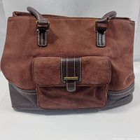 Front view of the handbag showing brown suede material with dark brown leather trims and dual handles. Includes front flap pocket with leather strap and brass-tone accent.