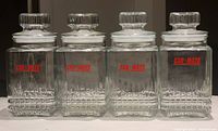 Set of four large clear glass canisters with lids, showing brand logo and textured square bases.
