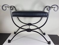 Front facing view of the vintage wrought iron bench with a black vinyl cushioned seat, showing curved scroll arms and legs.