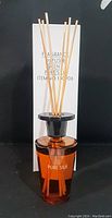 Fragrance diffuser bottle amber glass with black cap and natural reed sticks in front of white packaging box with product details.