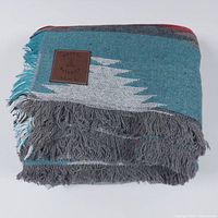 Folded multi-color woven beach blanket with fringed gray edges and leather branded patch.