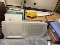 Four plastic storage bins, three with white lids, one folding bin in front, jumper cables on top of a cardboard box in the background.