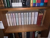 Shelf with multiple books including a uniform series with white spine, various titles visible, plus red hardcover book showing wear.