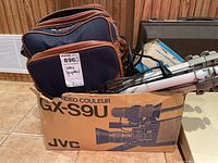 Wide view of lot showing JVC video camera box, camera bag, and tripod inside the box.