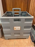 Front view of gray Gripngo collapsible cart with handle and wheels, placed on tile floor against wooden backdrop