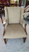 Front view of the beige upholstered armchair with wooden arms and legs, showing nail head trim and high wingback design.