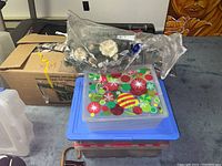 Multiple stacked plastic and cardboard boxes with Christmas decorations, including a sealed bag of white faux flowers, a container with a colorful holiday lid, and a brown cardboard box.