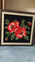 Full view of framed cross stitch art showing two red flowers with green leaves on black background.