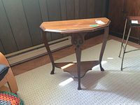 Full view of wooden demilune side table with three curved legs and lower shelf sitting on a carpeted floor.