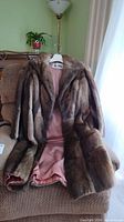 Brown mink fur coat hanging on a chair showing full front with pink satin lining partially visible.