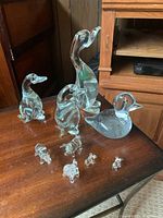 Nine clear glass animal figurines arranged on a wooden surface in front of brown furniture, showing different angles and lighting to highlight details.