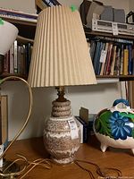 Full view of ceramic MCM table lamp with pleated cloth shade on wooden surface beside other items. Shows lamp design and general appearance.