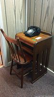 Photo showing black rotary phone sitting on small wood telephone table with wood chair beside it.