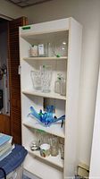 White laminate shelving unit in a room corner showing five shelves with glass items (not included) for scale.