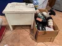 Wooden shoe shine box and cardboard box with shoe shining products such as polish cans, brushes and sponge on tiled floor.