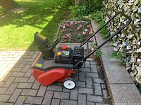 Side view of red and black Yard Machine snow blower on paved area next to garden with flowers. Shows front wheels and intake area.