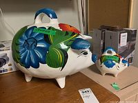 Photo showing both ceramic piggy banks positioned on a wooden surface with visible floral patterns and bright colors.
