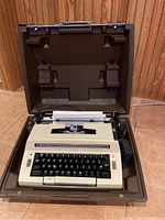 Smith-Corona electric portable typewriter inside an open brown hard case. The typewriter is beige with black keys and is positioned on a tiled floor with wood panel walls in the background.