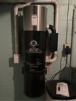 Full view of the ACV central vacuum unit installed on wall with attached pipes and accessories on side.