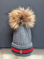 Front view of grey ribbed wool child’s hat with large tan coyote fur pompom and Gucci green and red striped cuff.