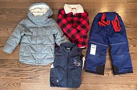 Lot of childrens winter clothing including jackets, vest, and snow pants on wood floor.