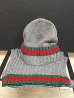 Front view of grey Gucci knit hat resting on folded matching scarf against black background.