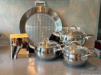 Overview showing three stainless steel pots with glass lids, large round perforated grilling dish, boxed Chicco coffee maker