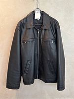 Full front view of black leather jacket hanging on hanger against light background. Shows zipper and pocket details.