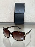 Front view of women's Prada sunglasses displaying brown gradient lenses and tortoiseshell frames, with the black Prada case behind.