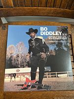 Front cover of the Bo Diddley 'Gunslinger' album showing Bo Diddley dressed as a cowboy with a red guitar on the ground.
