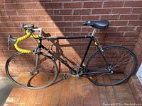 Side view of black road bike with yellow handlebar tape and clip-in pedals against brick wall.