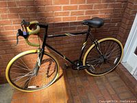 Full profile image of a black single speed fixie bike leaning against a brick wall featuring yellow handlebar tape and yellow tires
