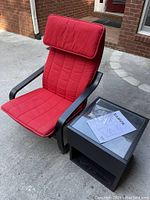Ikea Poang armchair with red cushions and black frame next to Ikea Ramvik black side table with glass top and instruction manual on top.