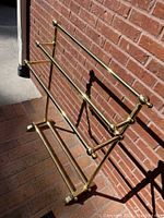 Brass hanging/drying rack leaning against a brick wall outside, showing three horizontal bars for hanging items and a rectangular base with four ball-footed legs for support.