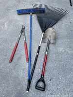 Photo showing four garden tools: a black metal leaf rake, a shovel with red D-handle, a blue push broom, and red-handled pruning loppers.
