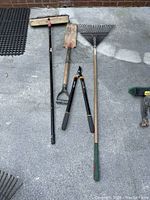 Photo of four garden tools laid out on concrete surface including a push broom, garden rake, pruner/lopper, and trench shovel.